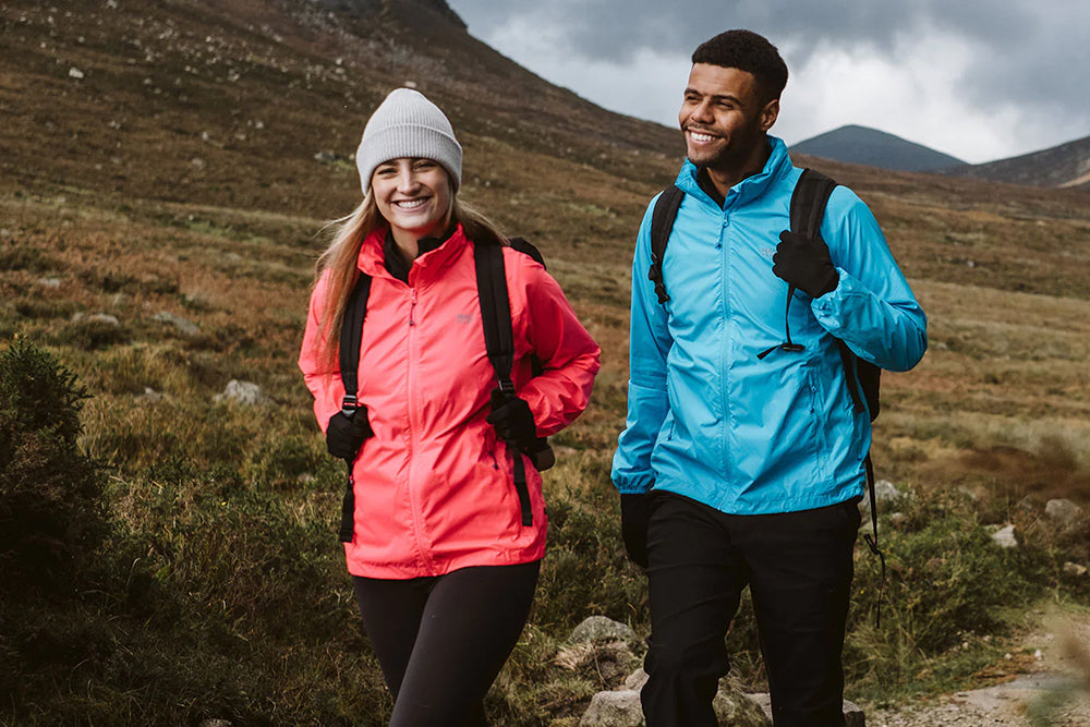 Two smiling hikers standing on a mountain trail, wearing colourful outdoor jackets and backpacks. 