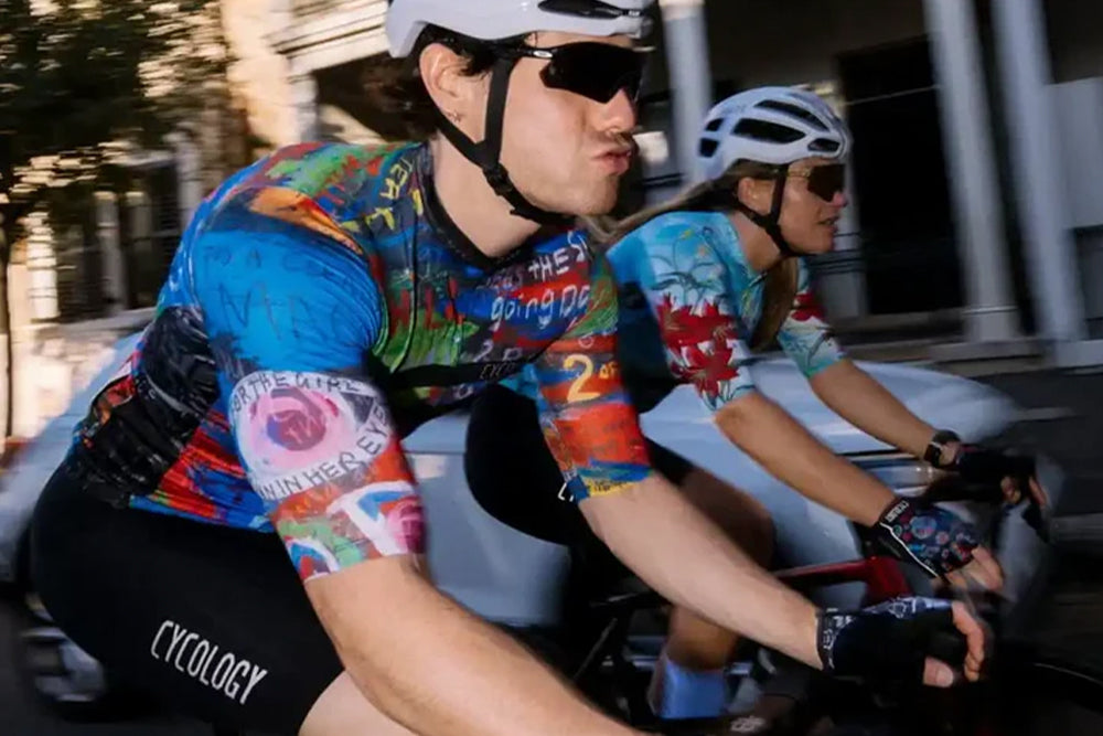 Two cyclists riding on a city street. The cyclist in the foreground is wearing an abstract patterned cycling jersey, black shorts with 'CYCLOGY' printed on the leg, and a white helmet.