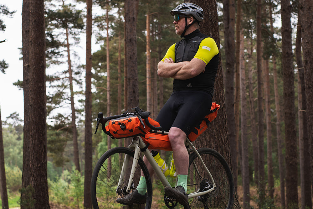 A cyclist wearing a black and neon yellow jersey, black shorts, and a helmet stands on a gravel bike with orange pannier bags in a dense pine forest.
