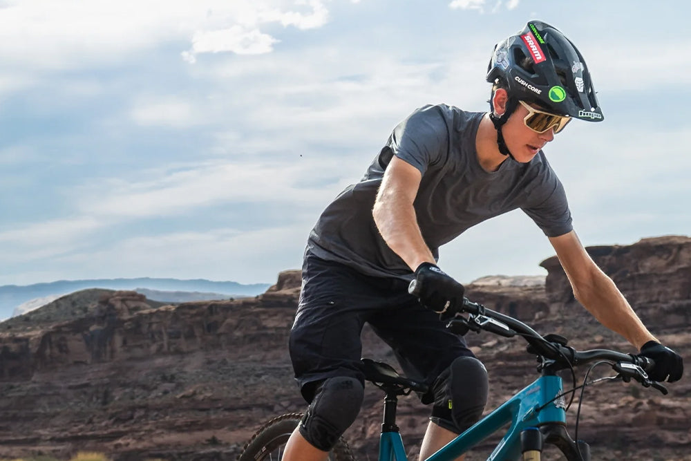 A mountain biker wearing a black helmet, sunglasses, a grey t-shirt, and black shorts rides a teal bike in a desert canyon landscape under a cloudy sky.