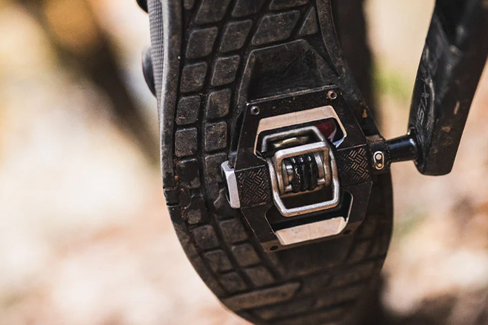 Close-up of a black bicycle pedal with a silver metal cleat attachment, shown against a blurred outdoor background.