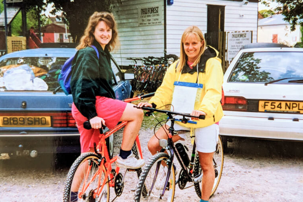 Two women with bicycles in a parking lot with vintage cars and a building in the background.