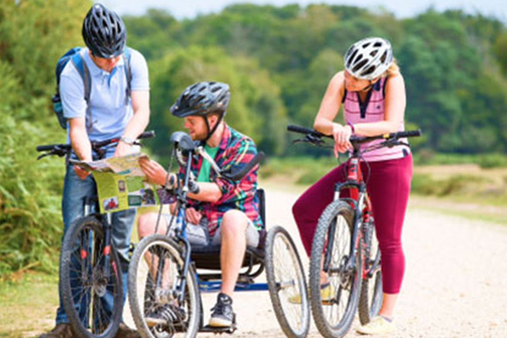 Three people on bicycles outdoors, two of whom are using adaptive bikes, with a scenic background.