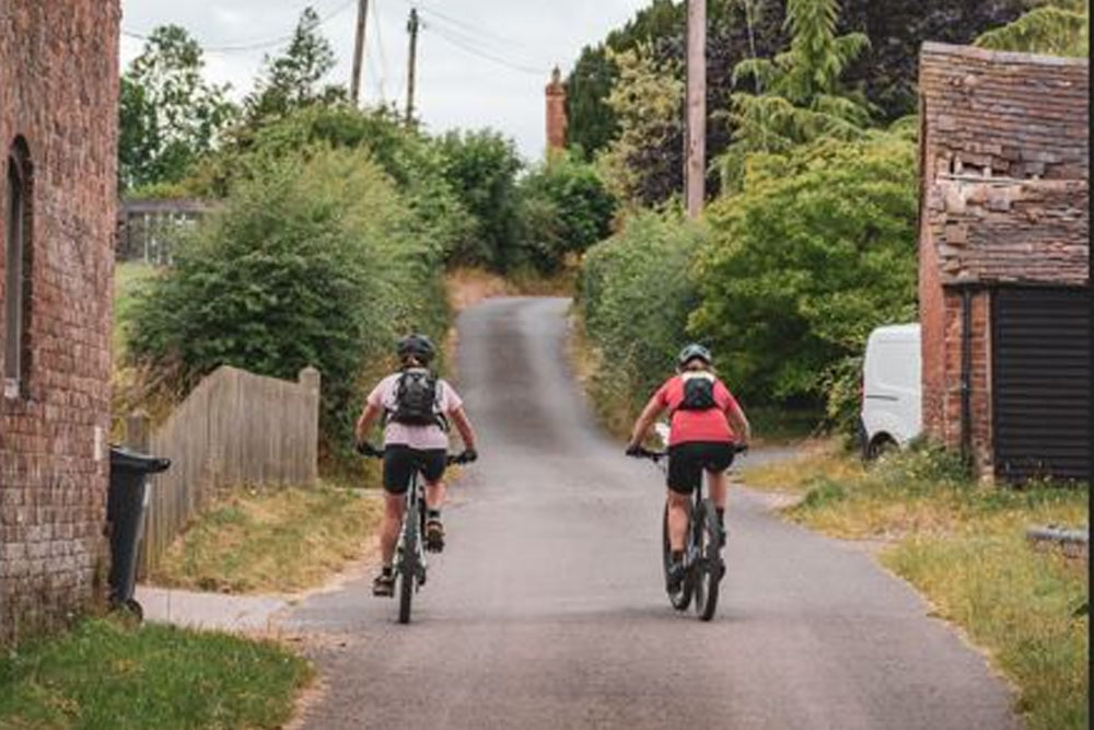 Two cyclists ride away on a narrow paved road between brick buildings and dense green shrubs, with utility poles and a white van visible in the background.