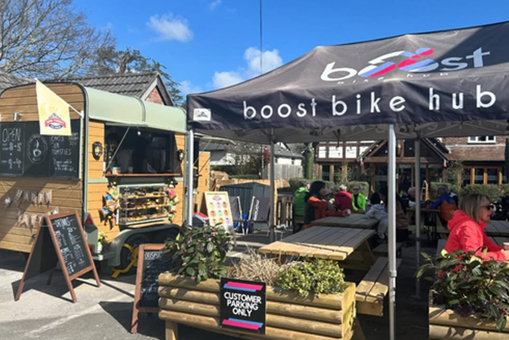 Outdoor cafe and bike hub scene with a black canopy tent printed 'boost bike hub' over wooden tables, a food truck, chalkboard signs, and seated people under a blue sky.