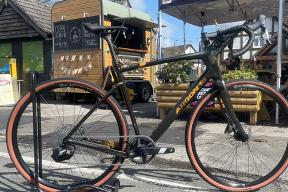 A black road/gravel bicycle with orange sidewall tires is displayed on a black stand in an outdoor street-side setting with a wooden food truck and storefront visible in the background.