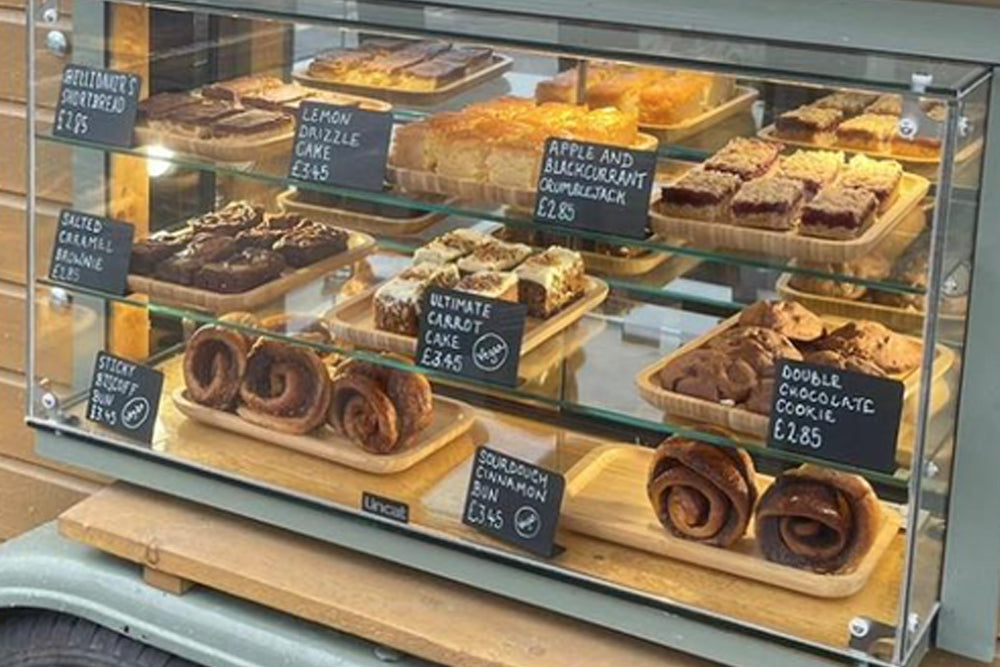 A glass bakery display case filled with assorted pastries and cakes with handwritten price tags on each item.