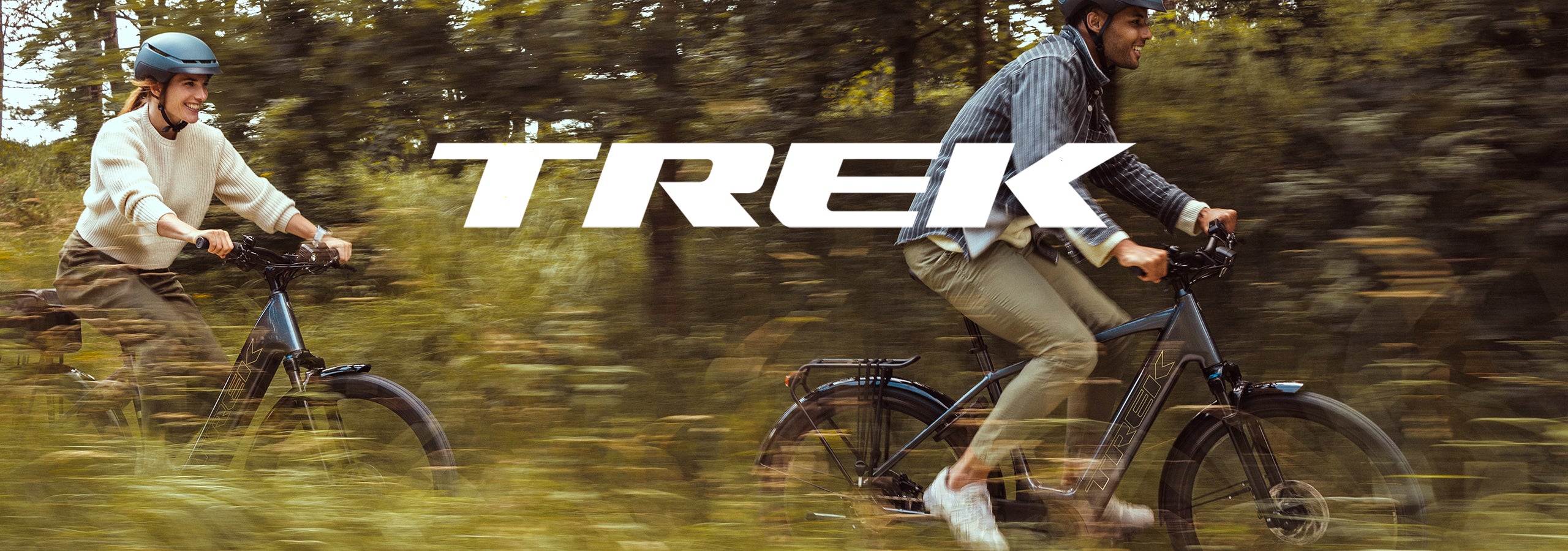 Two people riding electric bicycles on a blurred forest path, with the large TREK logo across the top center. The scene is outdoors with motion blur, and the bicycles appear dark blue/black.