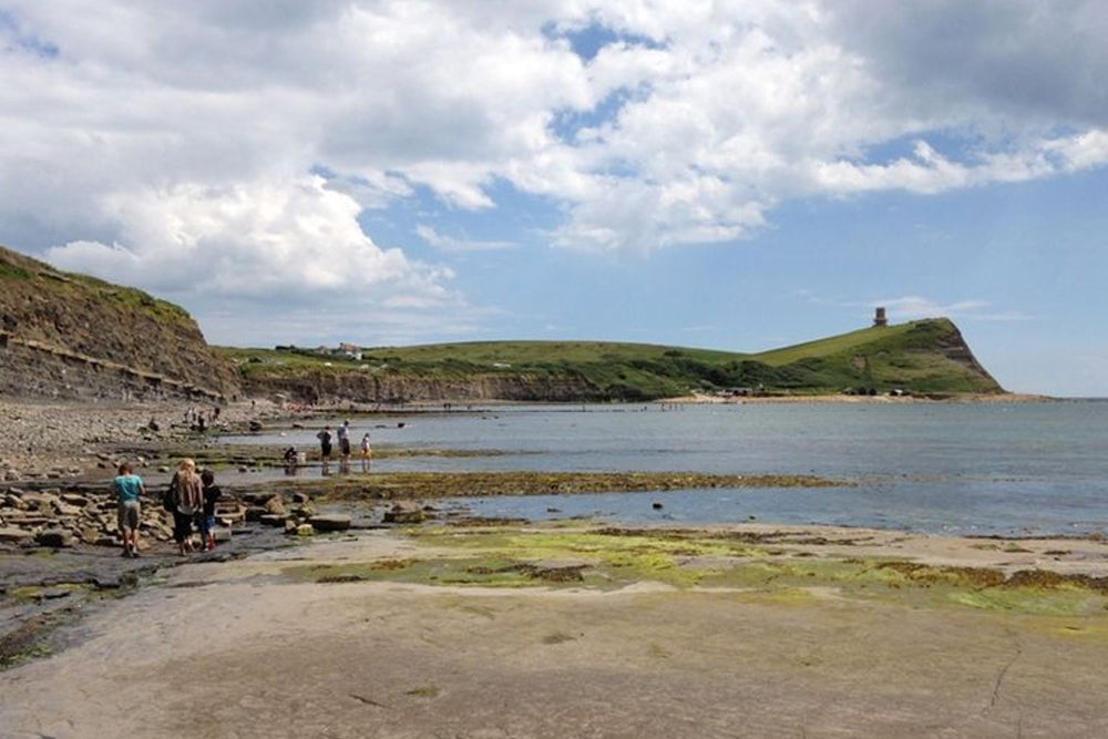 Coastal scene with people on a beach, cliffs, and a tower under a blue sky with clouds.