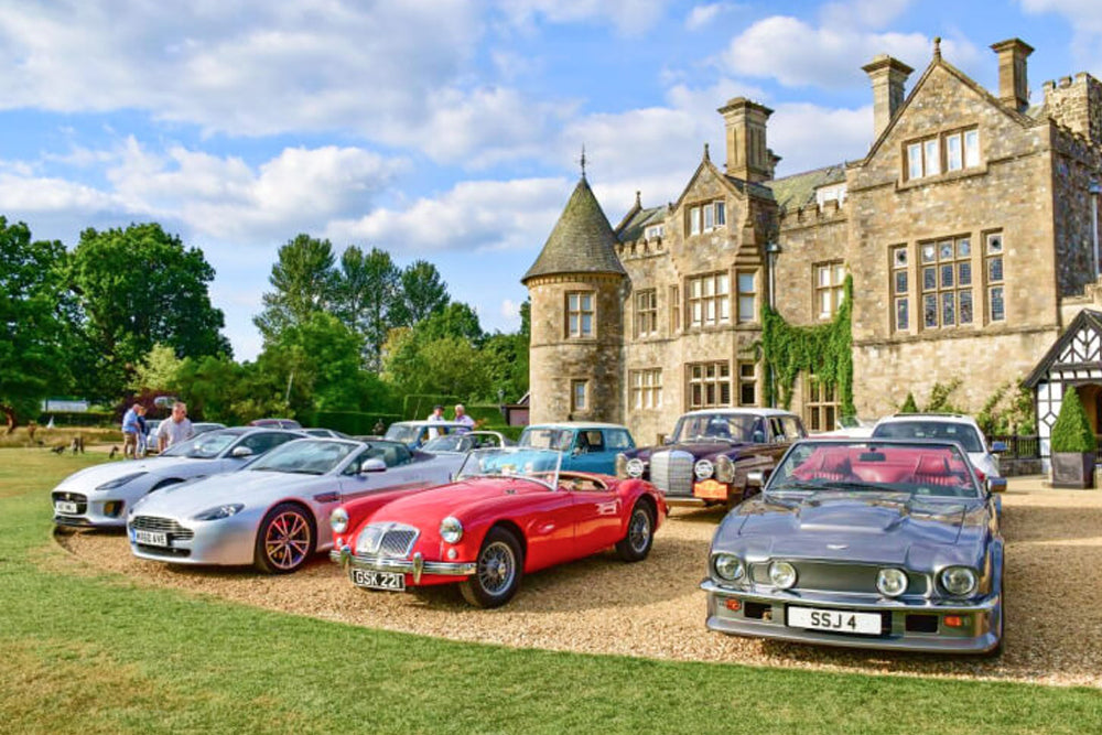 Collection of classic cars parked in front of a large stone building with greenery around.