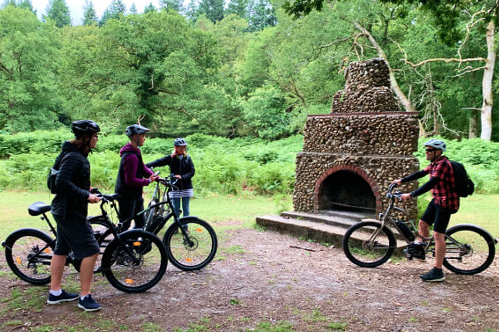Four people with electric bikes near a stone fireplace in a forest setting