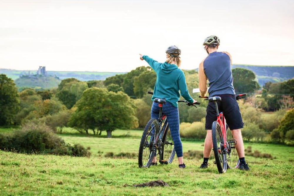 Two people with bicycles standing in a grassy field with trees and a clear sky in the background.