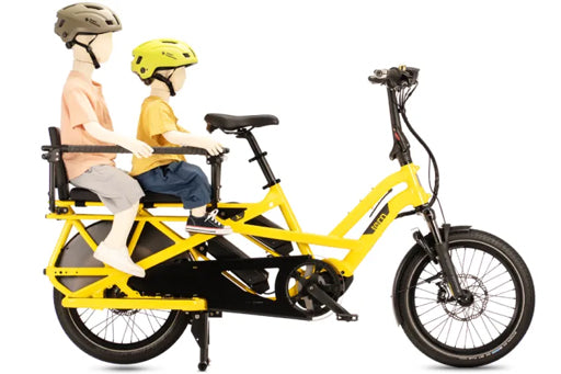 A bright yellow electric cargo bike with a rear passenger seat, shown on a white background with two child mannequins wearing helmets and seated on the bike.