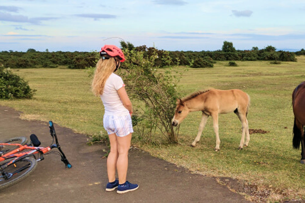 Girl with a bike and helmet standing next to a pony in a grassy field.