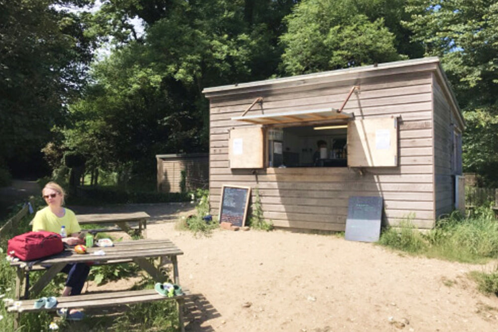 Person sitting at a picnic table in front of a small wooden building with open doors, surrounded by trees.