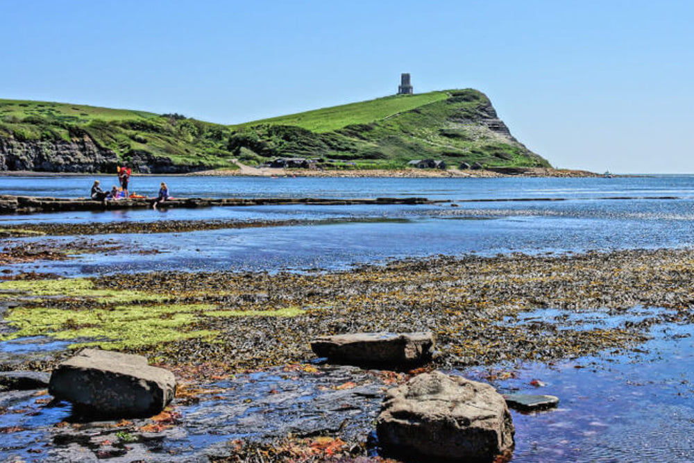 Beach scene with people on a rock, grassy hill, and tower in the distance.