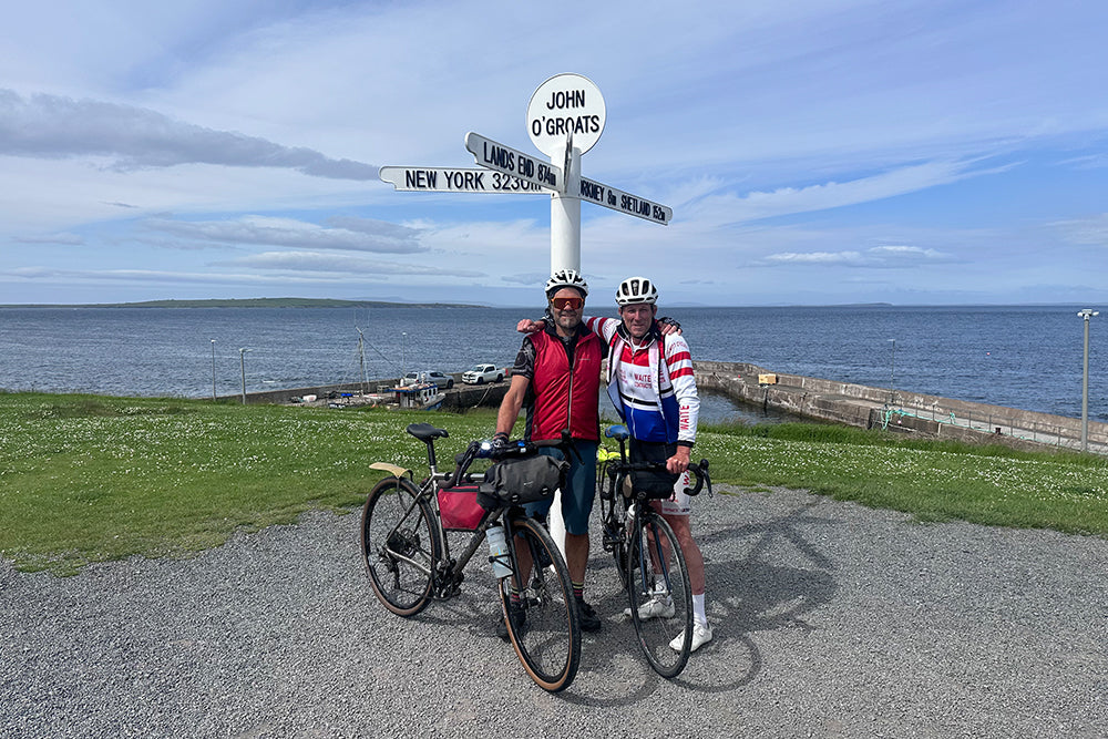 Two cyclists posing with their bikes in front of the John O'Groats signpost by the ocean, wearing cycling jerseys and helmets.