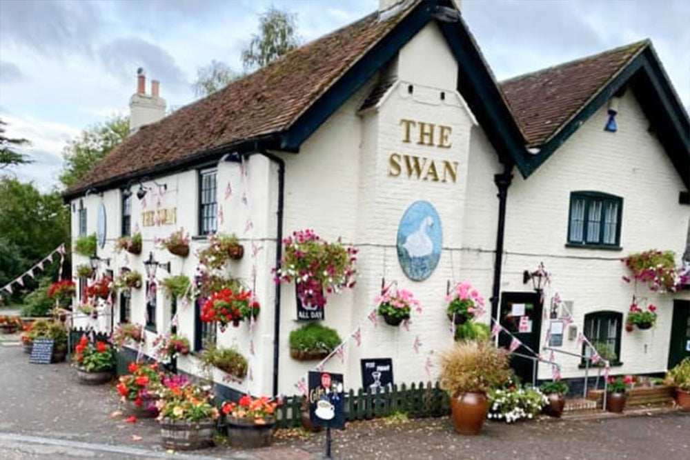 Pub named 'The Swan' with floral decorations on a sunny day.