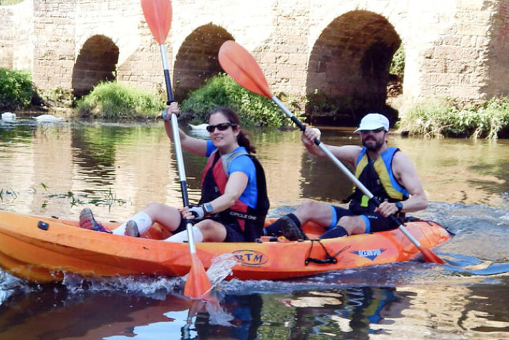 Two people paddling in an orange double kayak on a river in front of a stone bridge, wearing life jackets and holding orange paddles.