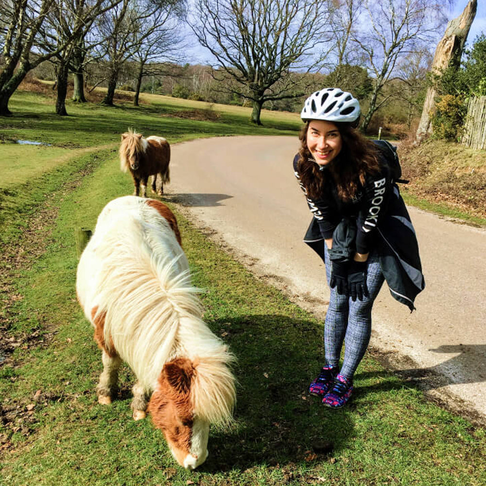 Woman in a helmet standing next to two small ponies on a path in a park.