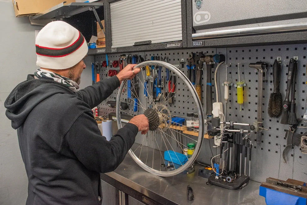 A man in a workshop wearing a white beanie with red and black stripes, working on a bicycle wheel mounted on a workbench. 